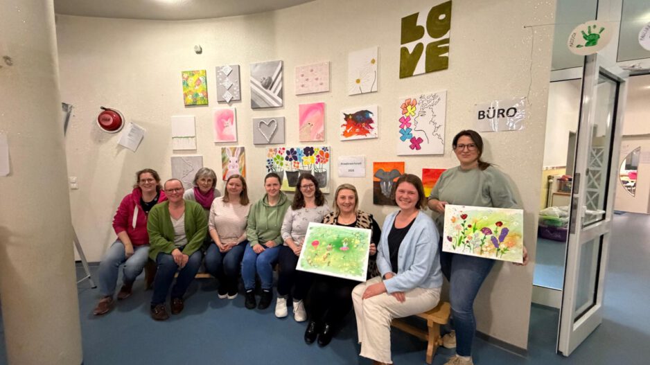 Gruppe von Frauen sitzt und steht in einem Raum vor einer Wand mit bunten Kunstwerken und hält selbst gestaltete Bilder aus einem Kreativworkshop in den Händen.