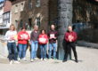 Sechs Personen stehen auf einem sonnigen Platz vor einem historischen Steingebäude und präsentieren Flyer sowie Broschüren vor einer kunstvoll gestalteten Säule.