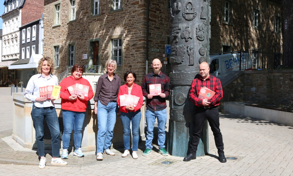 Sechs Personen stehen auf einem sonnigen Platz vor einem historischen Steingebäude und präsentieren Flyer sowie Broschüren vor einer kunstvoll gestalteten Säule.