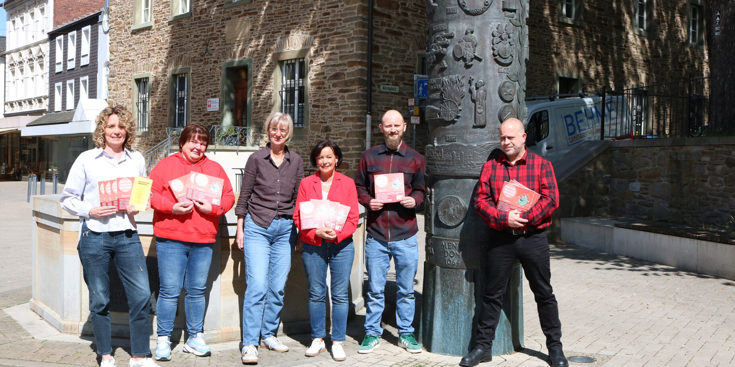 Sechs Personen stehen auf einem sonnigen Platz vor einem historischen Steingebäude und präsentieren Flyer sowie Broschüren vor einer kunstvoll gestalteten Säule.