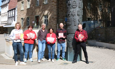 Sechs Personen stehen auf einem sonnigen Platz vor einem historischen Steingebäude und präsentieren Flyer sowie Broschüren vor einer kunstvoll gestalteten Säule.