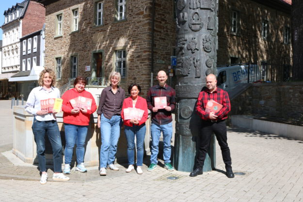 Sechs Personen stehen auf einem sonnigen Platz vor einem historischen Steingebäude und präsentieren Flyer sowie Broschüren vor einer kunstvoll gestalteten Säule.