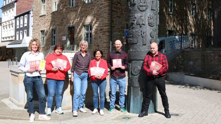 Sechs Personen stehen auf einem sonnigen Platz vor einem historischen Steingebäude und präsentieren Flyer sowie Broschüren vor einer kunstvoll gestalteten Säule.