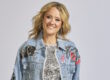 Eine lächelnde Frau mit schulterlangen blonden Haaren trägt ein glitzerndes Kleid und eine verzierte Jeansjacke vor neutralem Hintergrund.