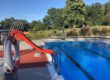 Ein Freibad mit einem Becken voll klarem Wasser und Schwimmbahnen; am Beckenrand steht eine rote Wasserrutsche, im Hintergrund sind Bäume und Liegewiesen zu sehen.