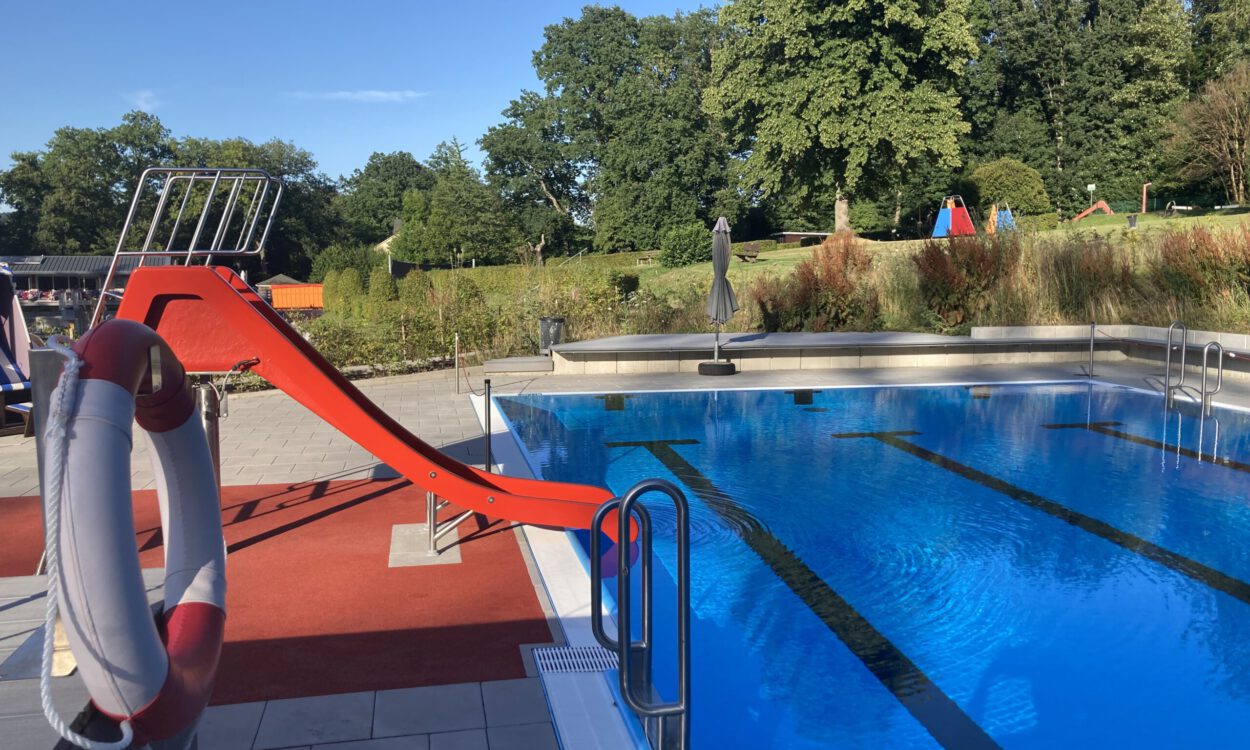 Ein Freibad mit einem Becken voll klarem Wasser und Schwimmbahnen; am Beckenrand steht eine rote Wasserrutsche, im Hintergrund sind Bäume und Liegewiesen zu sehen.