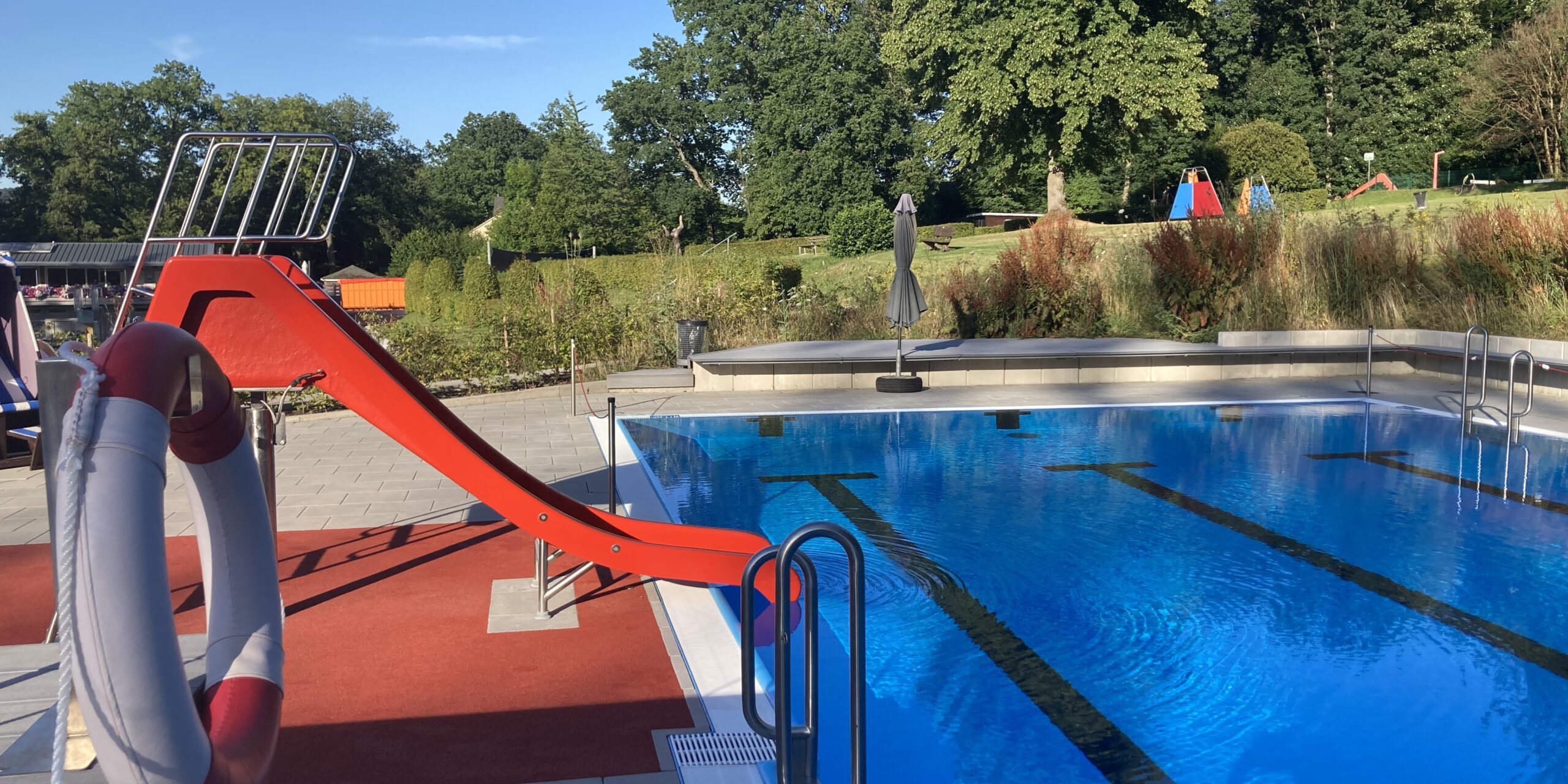 Ein Freibad mit einem Becken voll klarem Wasser und Schwimmbahnen; am Beckenrand steht eine rote Wasserrutsche, im Hintergrund sind Bäume und Liegewiesen zu sehen.