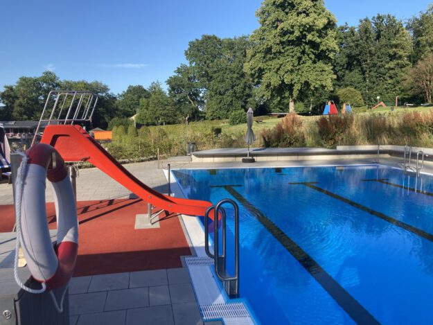 Ein Freibad mit einem Becken voll klarem Wasser und Schwimmbahnen; am Beckenrand steht eine rote Wasserrutsche, im Hintergrund sind Bäume und Liegewiesen zu sehen.