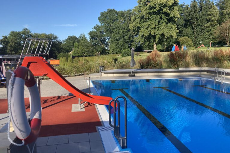Ein Freibad mit einem Becken voll klarem Wasser und Schwimmbahnen; am Beckenrand steht eine rote Wasserrutsche, im Hintergrund sind Bäume und Liegewiesen zu sehen.