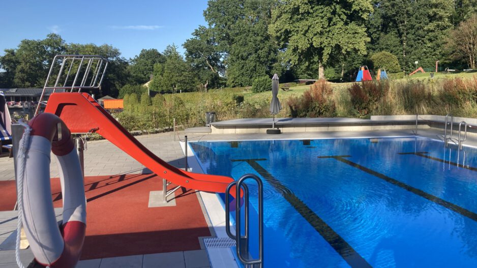 Ein Freibad mit einem Becken voll klarem Wasser und Schwimmbahnen; am Beckenrand steht eine rote Wasserrutsche, im Hintergrund sind Bäume und Liegewiesen zu sehen.