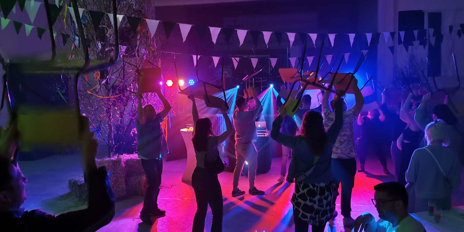 Menschen feiern in einer festlich beleuchteten Halle und tanzen mit hochgehobenen Stühlen unter buntem Licht, während Girlanden und Dekorationen für Partystimmung sorgen.