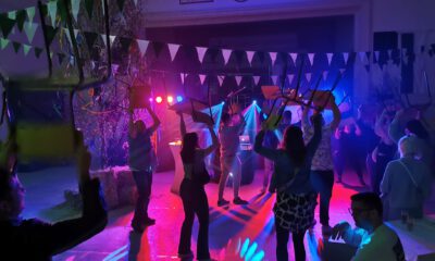 Menschen feiern in einer festlich beleuchteten Halle und tanzen mit hochgehobenen Stühlen unter buntem Licht, während Girlanden und Dekorationen für Partystimmung sorgen.