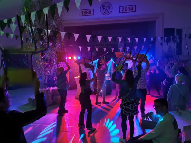 Menschen feiern in einer festlich beleuchteten Halle und tanzen mit hochgehobenen Stühlen unter buntem Licht, während Girlanden und Dekorationen für Partystimmung sorgen.