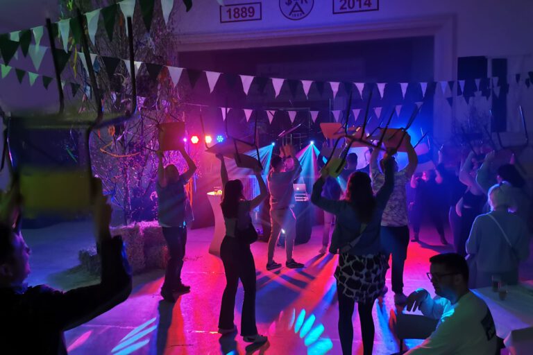 Menschen feiern in einer festlich beleuchteten Halle und tanzen mit hochgehobenen Stühlen unter buntem Licht, während Girlanden und Dekorationen für Partystimmung sorgen.