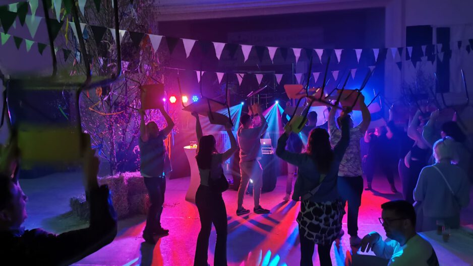 Menschen feiern in einer festlich beleuchteten Halle und tanzen mit hochgehobenen Stühlen unter buntem Licht, während Girlanden und Dekorationen für Partystimmung sorgen.