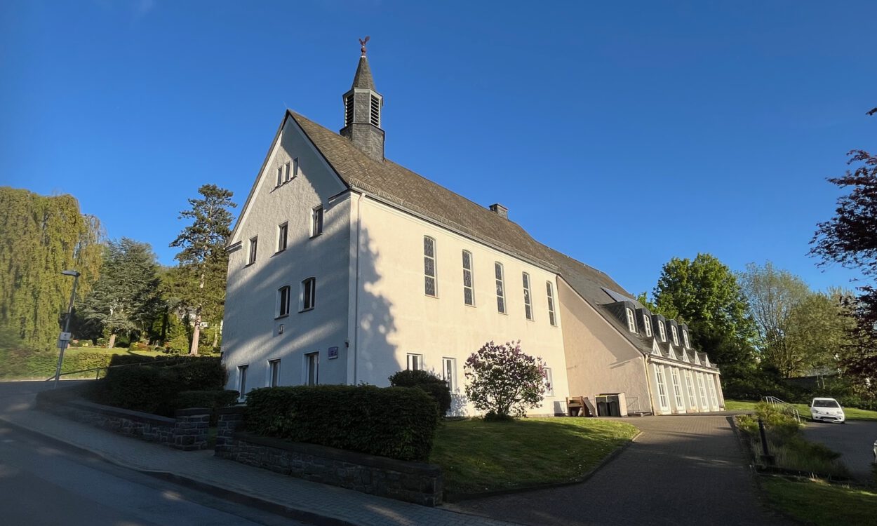 Ein großes helles Kirchengebäude mit kleinem Dachreiter steht an einer ruhigen Straße, umgeben von Grünflächen und Bäumen unter blauem Himmel.