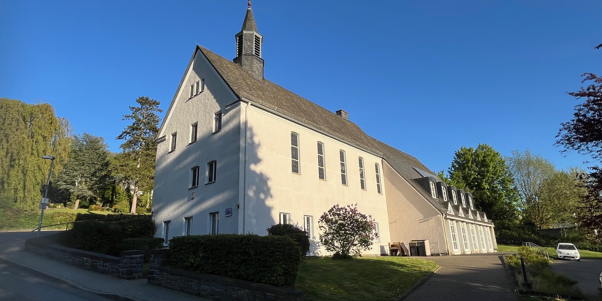 Ein großes helles Kirchengebäude mit kleinem Dachreiter steht an einer ruhigen Straße, umgeben von Grünflächen und Bäumen unter blauem Himmel.