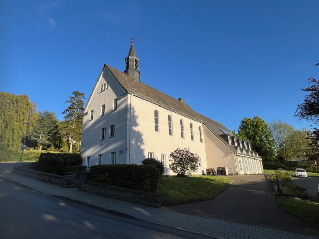 Ein großes helles Kirchengebäude mit kleinem Dachreiter steht an einer ruhigen Straße, umgeben von Grünflächen und Bäumen unter blauem Himmel.