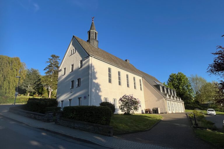 Ein großes helles Kirchengebäude mit kleinem Dachreiter steht an einer ruhigen Straße, umgeben von Grünflächen und Bäumen unter blauem Himmel.