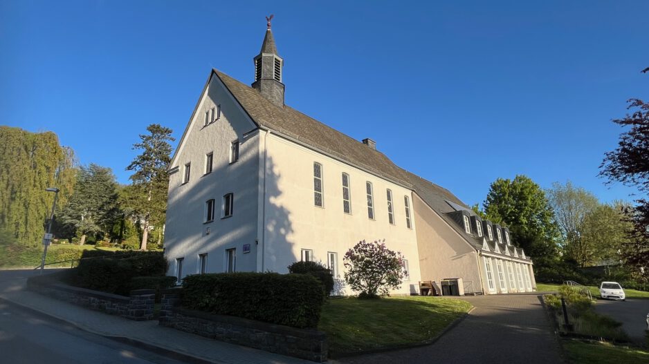 Ein großes helles Kirchengebäude mit kleinem Dachreiter steht an einer ruhigen Straße, umgeben von Grünflächen und Bäumen unter blauem Himmel.