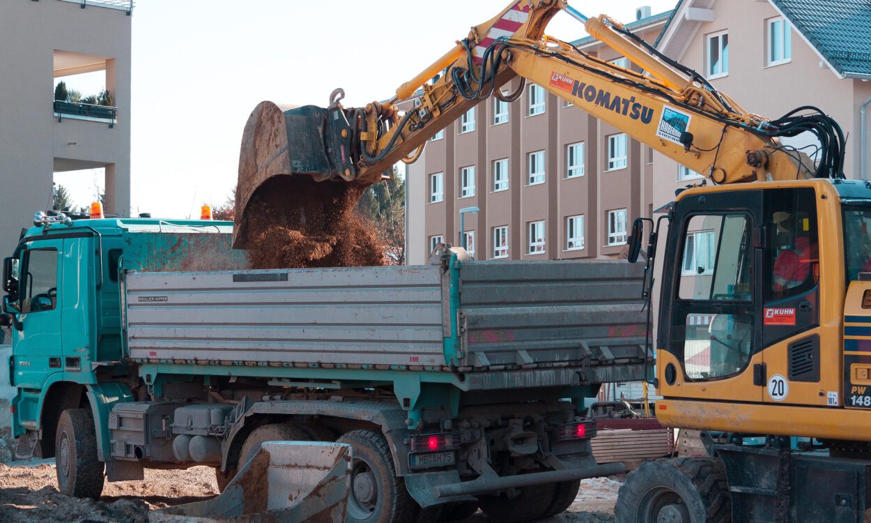 Ein Bagger lädt auf einer Baustelle Erde in die Ladefläche eines Lastwagens; im Hintergrund stehen mehrstöckige Wohnhäuser.