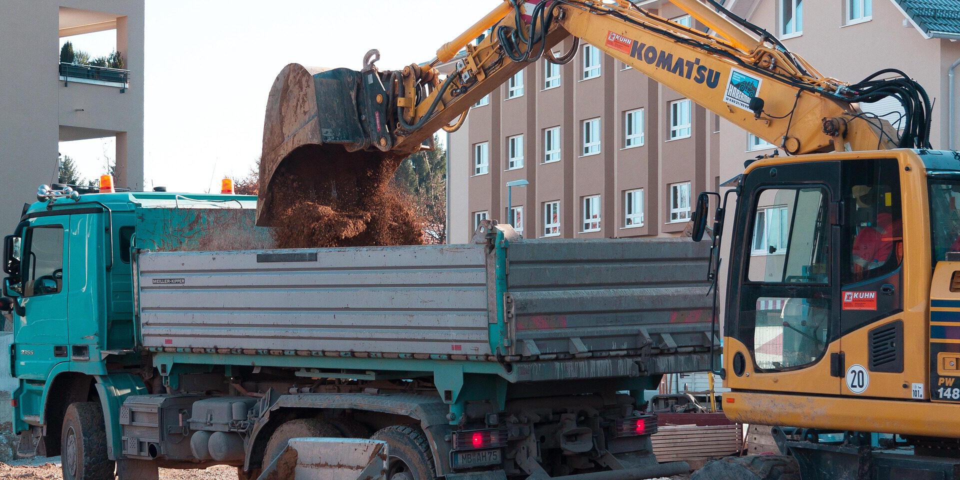 Ein Bagger lädt auf einer Baustelle Erde in die Ladefläche eines Lastwagens; im Hintergrund stehen mehrstöckige Wohnhäuser.