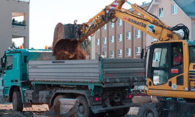 Ein Bagger lädt auf einer Baustelle Erde in die Ladefläche eines Lastwagens; im Hintergrund stehen mehrstöckige Wohnhäuser.