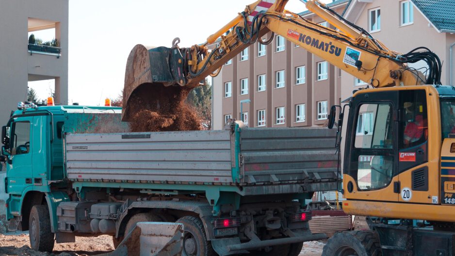 Ein Bagger lädt auf einer Baustelle Erde in die Ladefläche eines Lastwagens; im Hintergrund stehen mehrstöckige Wohnhäuser.