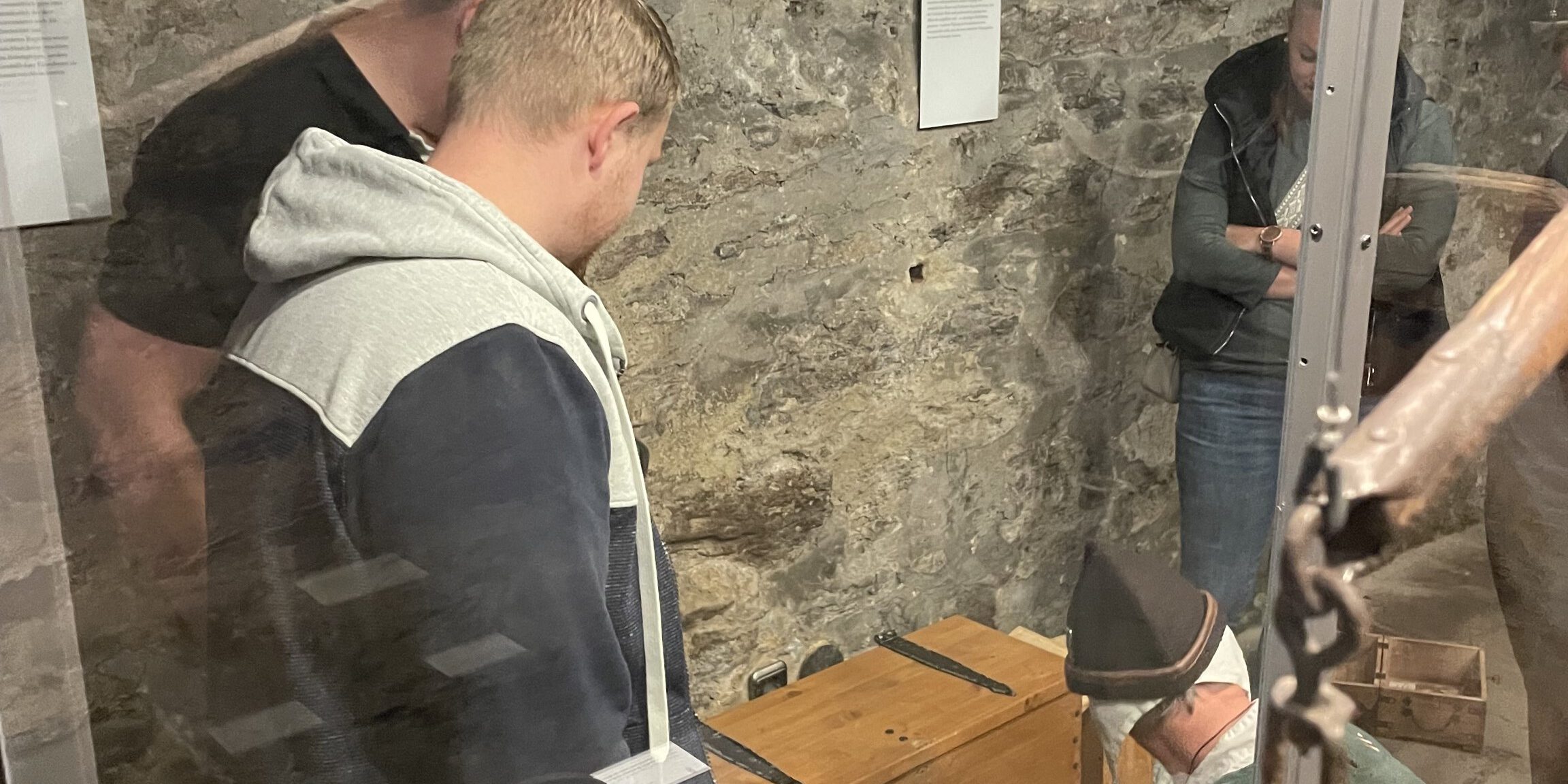Mehrere Menschen betrachten in einem historischen Kellerraum eine Ausstellung mit alten Gegenständen hinter Glasvitrinen und Steinmauern im Hintergrund.