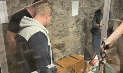 Mehrere Menschen betrachten in einem historischen Kellerraum eine Ausstellung mit alten Gegenständen hinter Glasvitrinen und Steinmauern im Hintergrund.