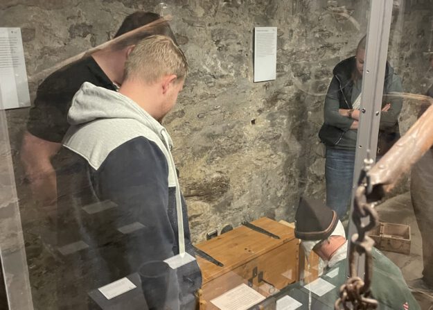 Mehrere Menschen betrachten in einem historischen Kellerraum eine Ausstellung mit alten Gegenständen hinter Glasvitrinen und Steinmauern im Hintergrund.