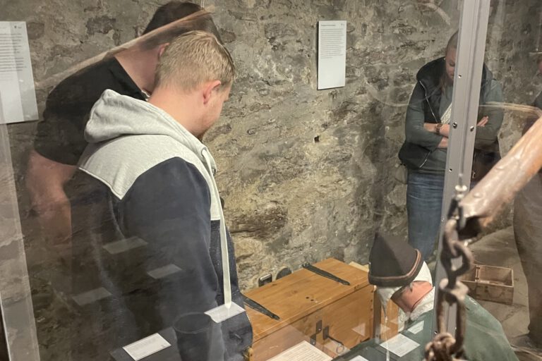 Mehrere Menschen betrachten in einem historischen Kellerraum eine Ausstellung mit alten Gegenständen hinter Glasvitrinen und Steinmauern im Hintergrund.