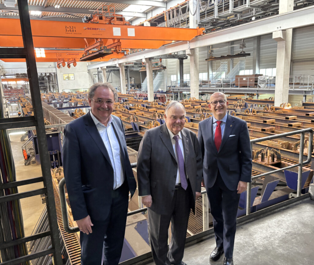 Drei Männer in Anzügen stehen in einer großen Industriehalle vor einer Produktionsanlage mit vielen Metallformen und einem orangefarbenen Hallenkran im Hintergrund.