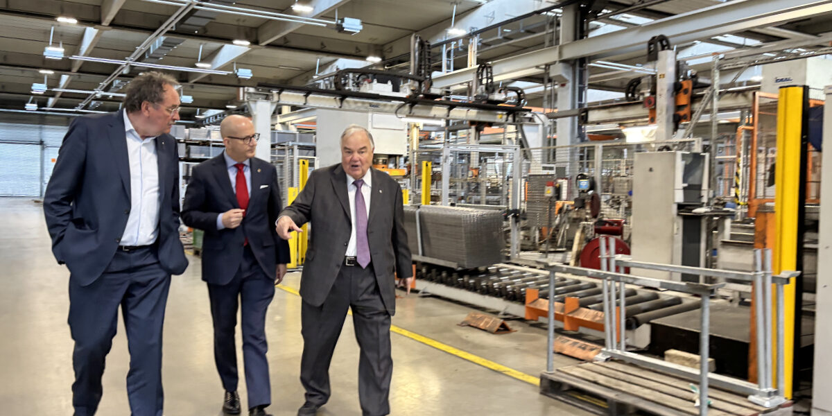 Drei Männer in Anzügen gehen durch eine Industriehalle und unterhalten sich, während sie an Maschinen und Förderanlagen vorbeilaufen; einer zeigt dabei nach vorne.