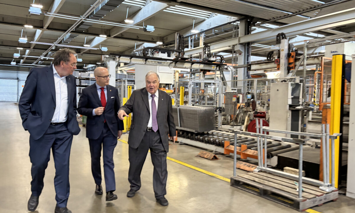 Drei Männer in Anzügen gehen durch eine Industriehalle und unterhalten sich, während sie an Maschinen und Förderanlagen vorbeilaufen; einer zeigt dabei nach vorne.