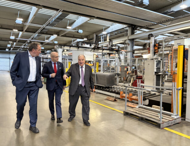 Drei Männer in Anzügen gehen durch eine Industriehalle und unterhalten sich, während sie an Maschinen und Förderanlagen vorbeilaufen; einer zeigt dabei nach vorne.