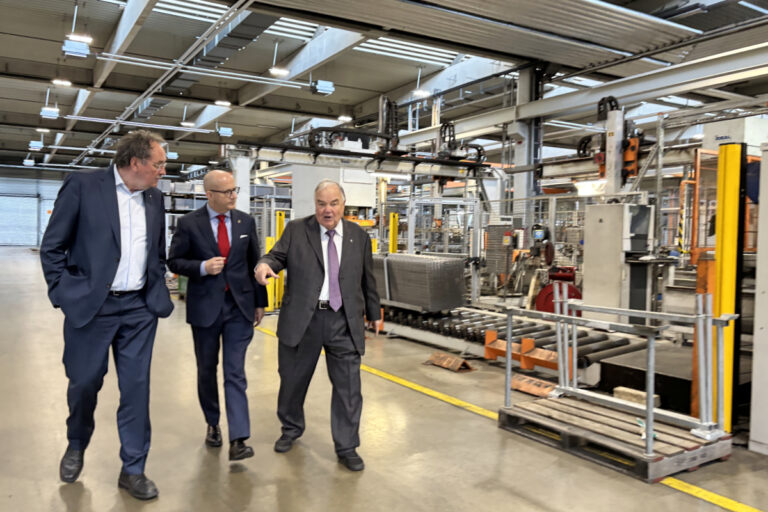 Drei Männer in Anzügen gehen durch eine Industriehalle und unterhalten sich, während sie an Maschinen und Förderanlagen vorbeilaufen; einer zeigt dabei nach vorne.