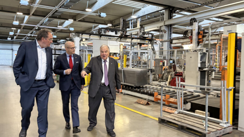 Drei Männer in Anzügen gehen durch eine Industriehalle und unterhalten sich, während sie an Maschinen und Förderanlagen vorbeilaufen; einer zeigt dabei nach vorne.