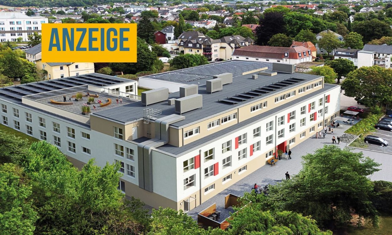 Luftaufnahme einer modernen Wohnanlage mit mehreren Gebäudeteilen, begrünten Außenflächen, Parkplätzen und einer Dachterrasse inmitten eines Wohngebiets. Oben links steht ein Hinweis, dass es sich um eine Anzeige handelt.
