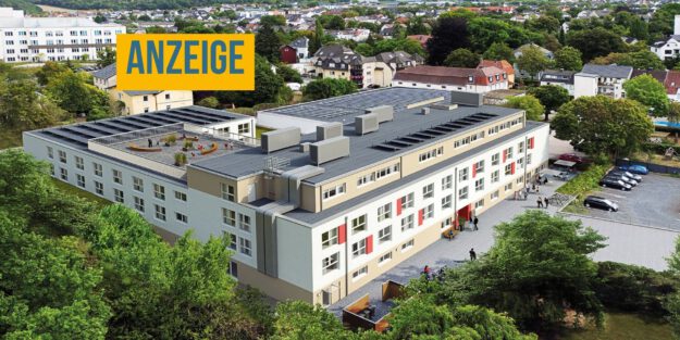 Luftaufnahme einer modernen Wohnanlage mit mehreren Gebäudeteilen, begrünten Außenflächen, Parkplätzen und einer Dachterrasse inmitten eines Wohngebiets. Oben links steht ein Hinweis, dass es sich um eine Anzeige handelt.