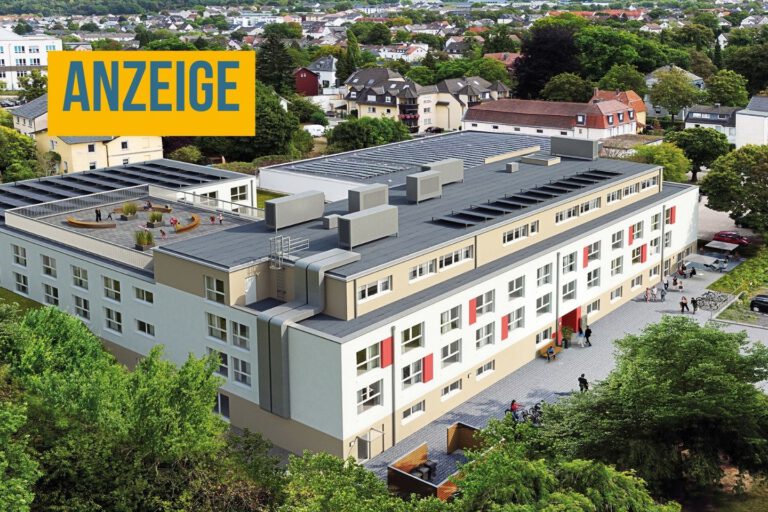 Luftaufnahme einer modernen Wohnanlage mit mehreren Gebäudeteilen, begrünten Außenflächen, Parkplätzen und einer Dachterrasse inmitten eines Wohngebiets. Oben links steht ein Hinweis, dass es sich um eine Anzeige handelt.