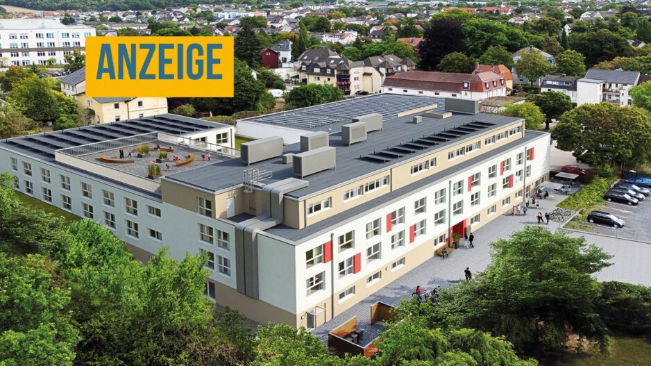 Luftaufnahme einer modernen Wohnanlage mit mehreren Gebäudeteilen, begrünten Außenflächen, Parkplätzen und einer Dachterrasse inmitten eines Wohngebiets. Oben links steht ein Hinweis, dass es sich um eine Anzeige handelt.
