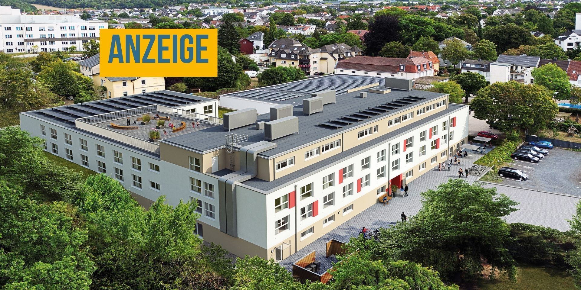 Luftaufnahme einer modernen Wohnanlage mit mehreren Gebäudeteilen, begrünten Außenflächen, Parkplätzen und einer Dachterrasse inmitten eines Wohngebiets. Oben links steht ein Hinweis, dass es sich um eine Anzeige handelt.