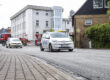 Ein weißes Elektroauto fährt auf einer frisch mit Splitt bearbeiteten Straße durch ein Stadtgebiet; im Hintergrund sind weitere Fahrzeuge, Baustellenabsperrungen und Gebäude zu sehen.