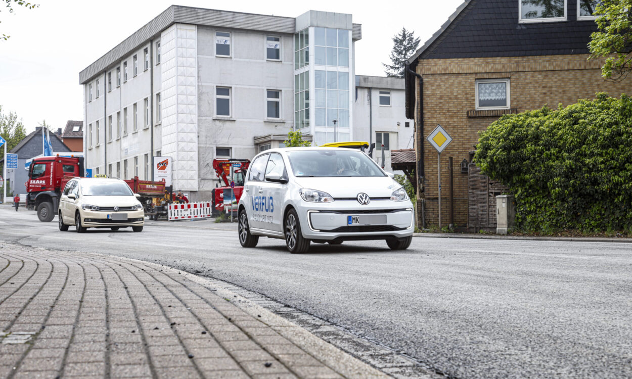 Ein weißes Elektroauto fährt auf einer frisch mit Splitt bearbeiteten Straße durch ein Stadtgebiet; im Hintergrund sind weitere Fahrzeuge, Baustellenabsperrungen und Gebäude zu sehen.