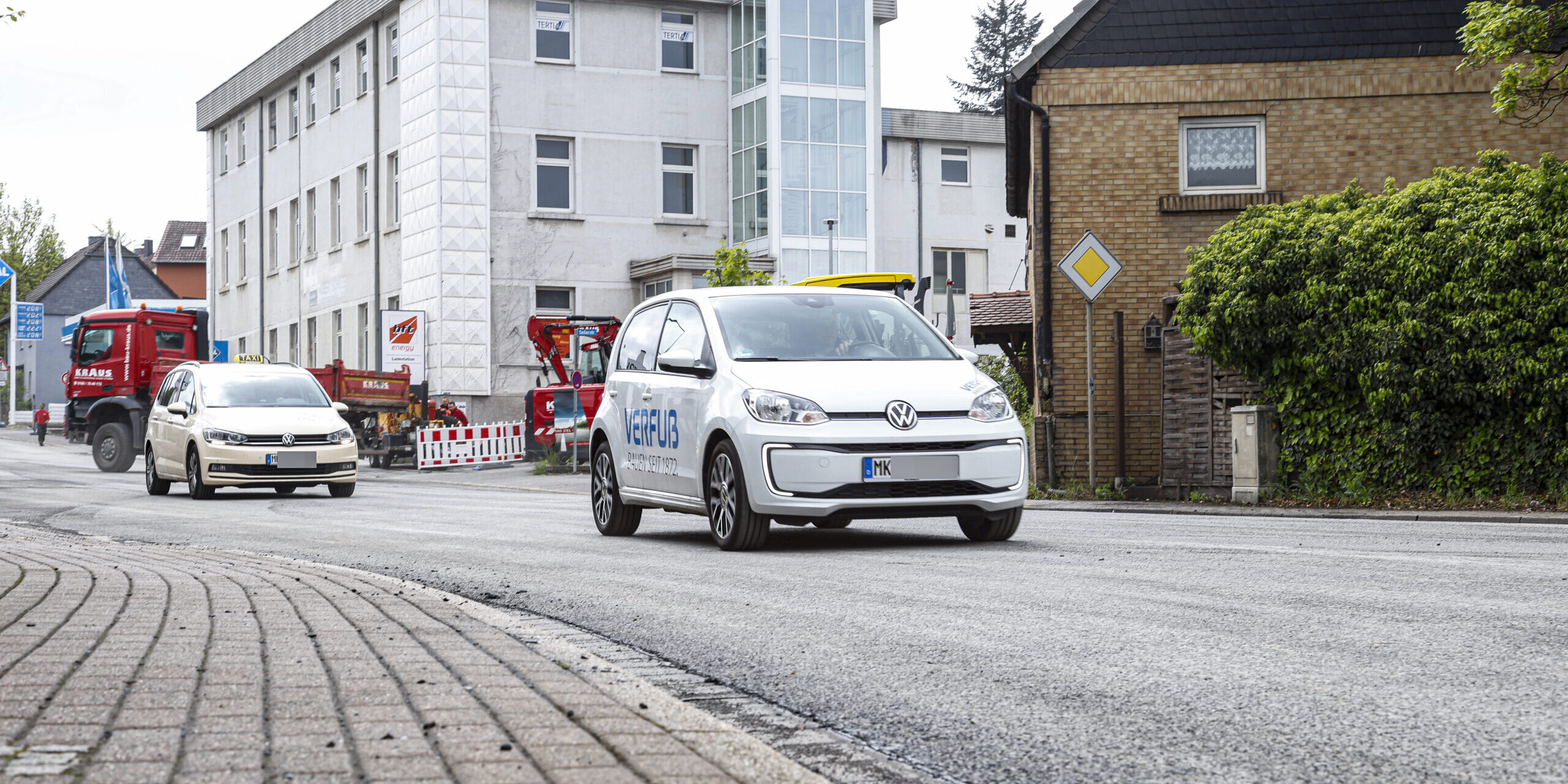 Ein weißes Elektroauto fährt auf einer frisch mit Splitt bearbeiteten Straße durch ein Stadtgebiet; im Hintergrund sind weitere Fahrzeuge, Baustellenabsperrungen und Gebäude zu sehen.