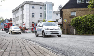 Ein weißes Elektroauto fährt auf einer frisch mit Splitt bearbeiteten Straße durch ein Stadtgebiet; im Hintergrund sind weitere Fahrzeuge, Baustellenabsperrungen und Gebäude zu sehen.