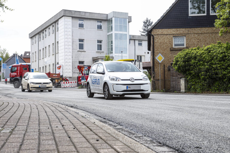 Ein weißes Elektroauto fährt auf einer frisch mit Splitt bearbeiteten Straße durch ein Stadtgebiet; im Hintergrund sind weitere Fahrzeuge, Baustellenabsperrungen und Gebäude zu sehen.