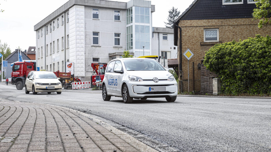 Ein weißes Elektroauto fährt auf einer frisch mit Splitt bearbeiteten Straße durch ein Stadtgebiet; im Hintergrund sind weitere Fahrzeuge, Baustellenabsperrungen und Gebäude zu sehen.