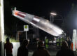 Bei Nacht wird ein großes Rotorblatt einer Windkraftanlage mit einem Kran über eine Straße transportiert, während mehrere Menschen und Einsatzkräfte den Transport beobachten.